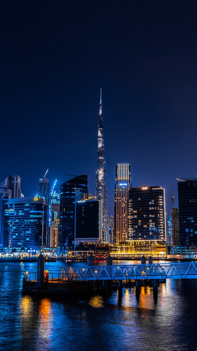 A city at night with lots of blue lights on the buildings reflecting on a mostly calm lake. One building is mostly orange.